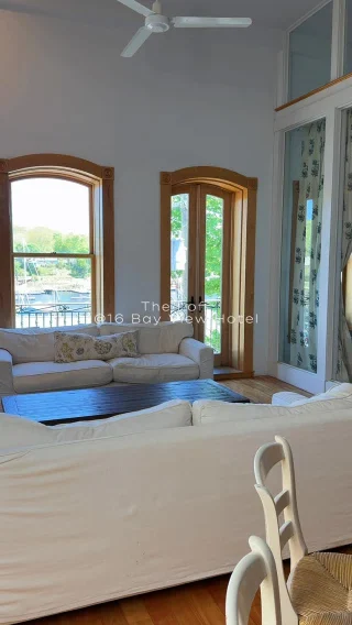 A cozy seating area with a white couch, wooden tables, and arch windows overlooking a scenic view.